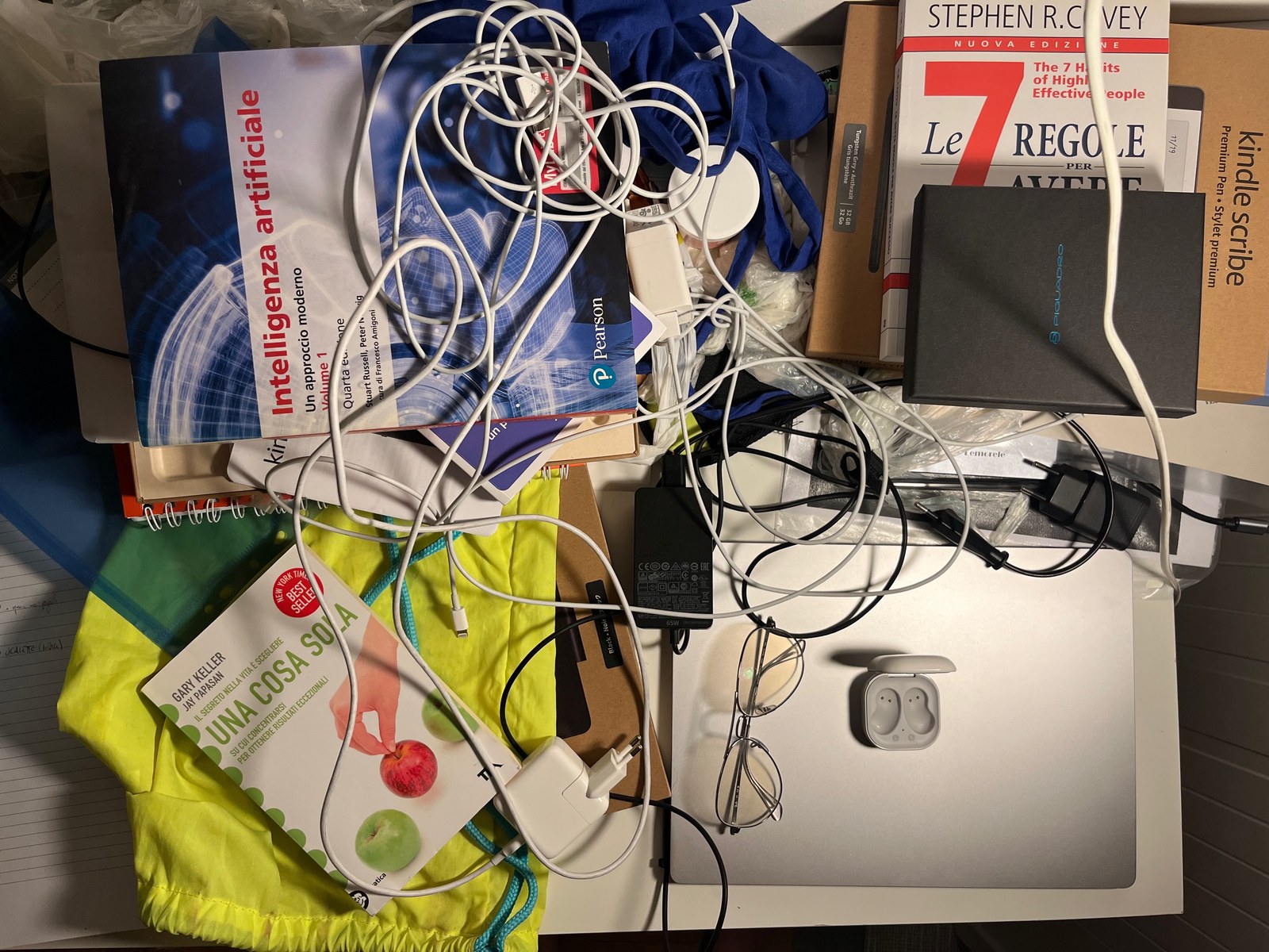 Scrivania di casa in disordine la sera, vista dall'alto: cavi bianchi aggrovigliati, caricabatterie, un laptop chiuso, occhiali, una custodia di auricolari aperta e vuota, e i libri Intelligenza artificiale di Russell e Norvig, Le 7 regole per avere successo di Stephen Covey e Una cosa sola di Gary Keller